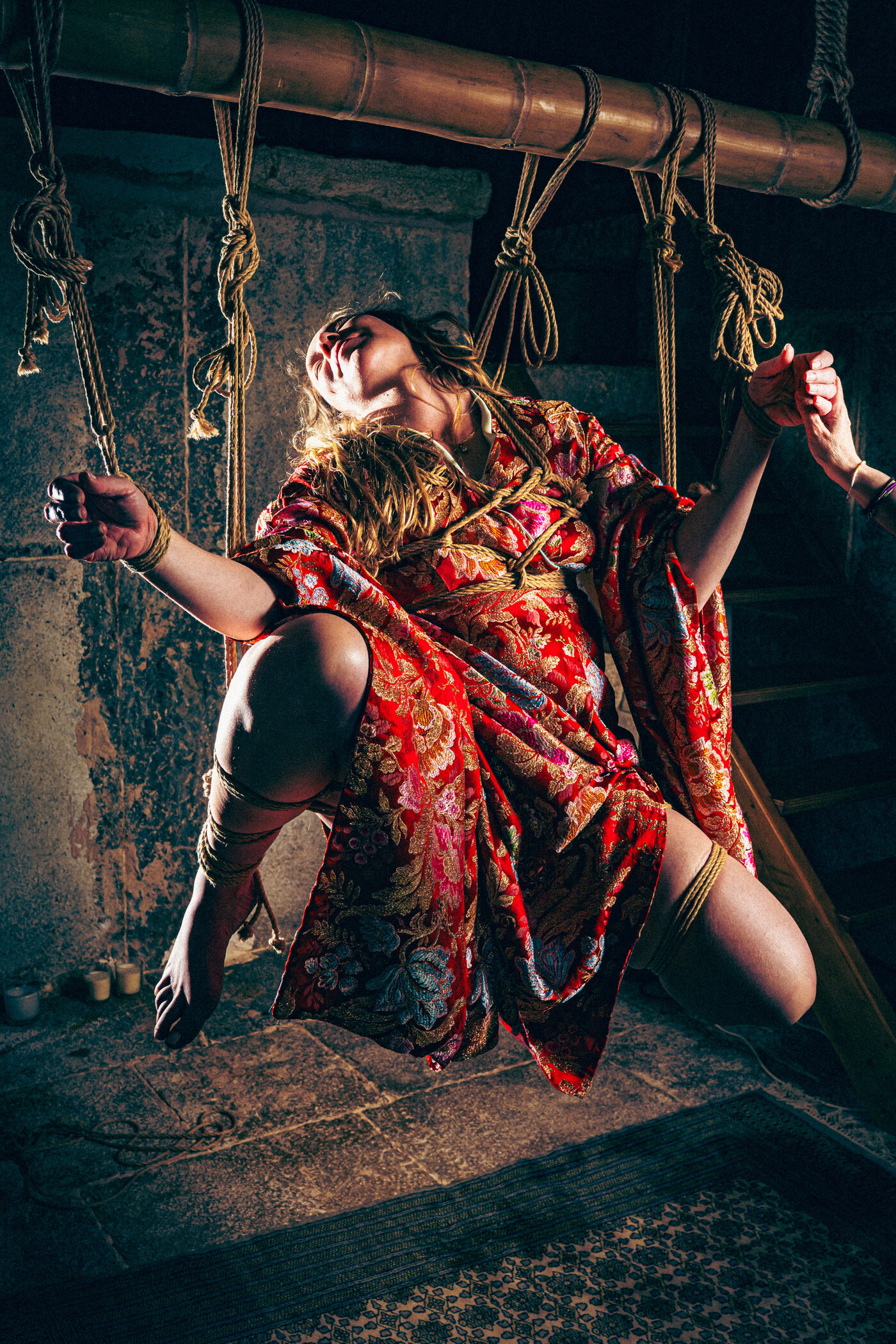 Suspension in a red floral kimono from a bamboo beam; a hand reaches in to hold the model's bound hand